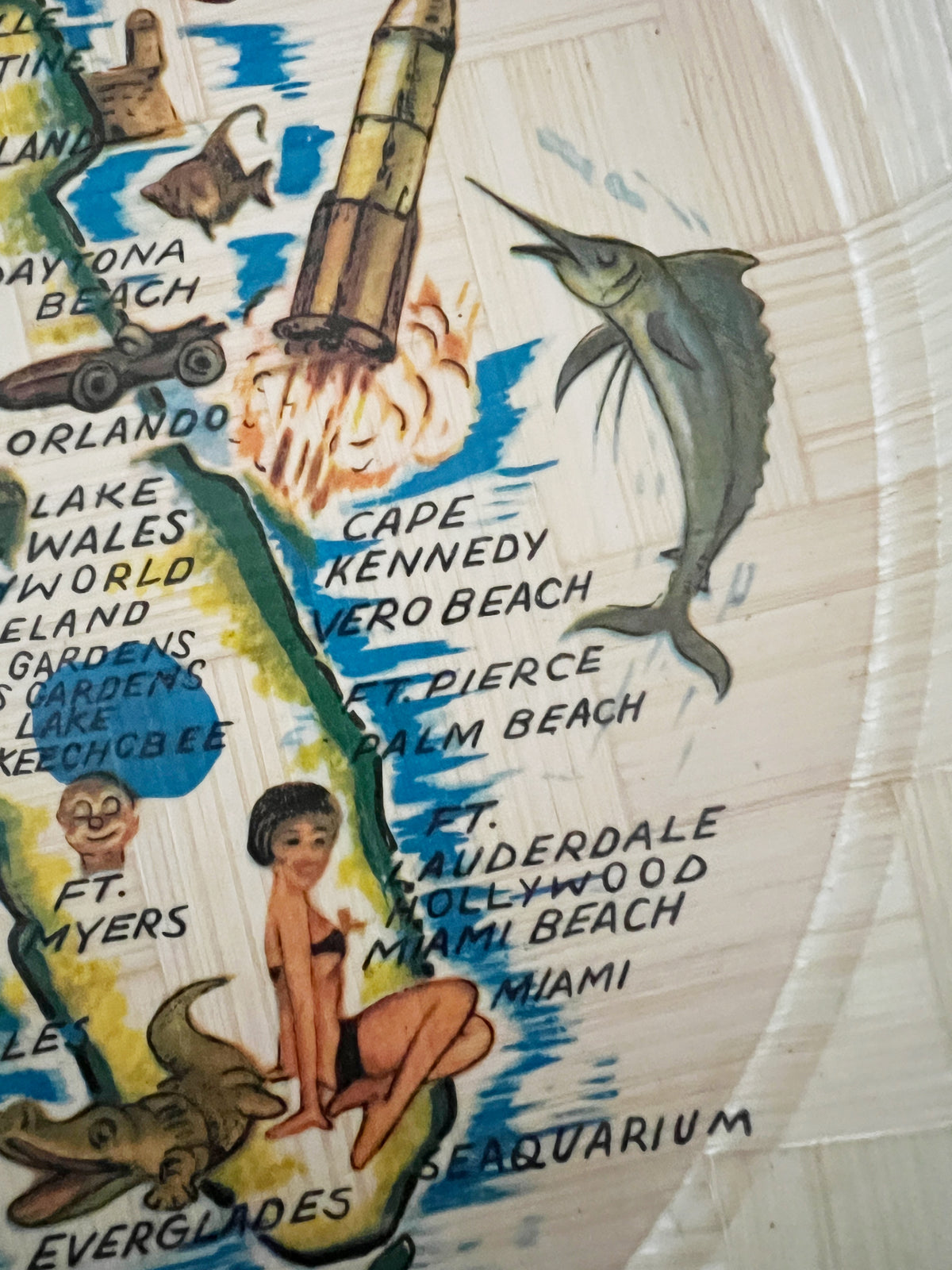 A close-up of a vintage-style illustrated map print featuring Florida landmarks and names, including Cape Kennedy, Miami Beach, and Everglades, with hand-drawn figures and a marlin on a cream-colored background.